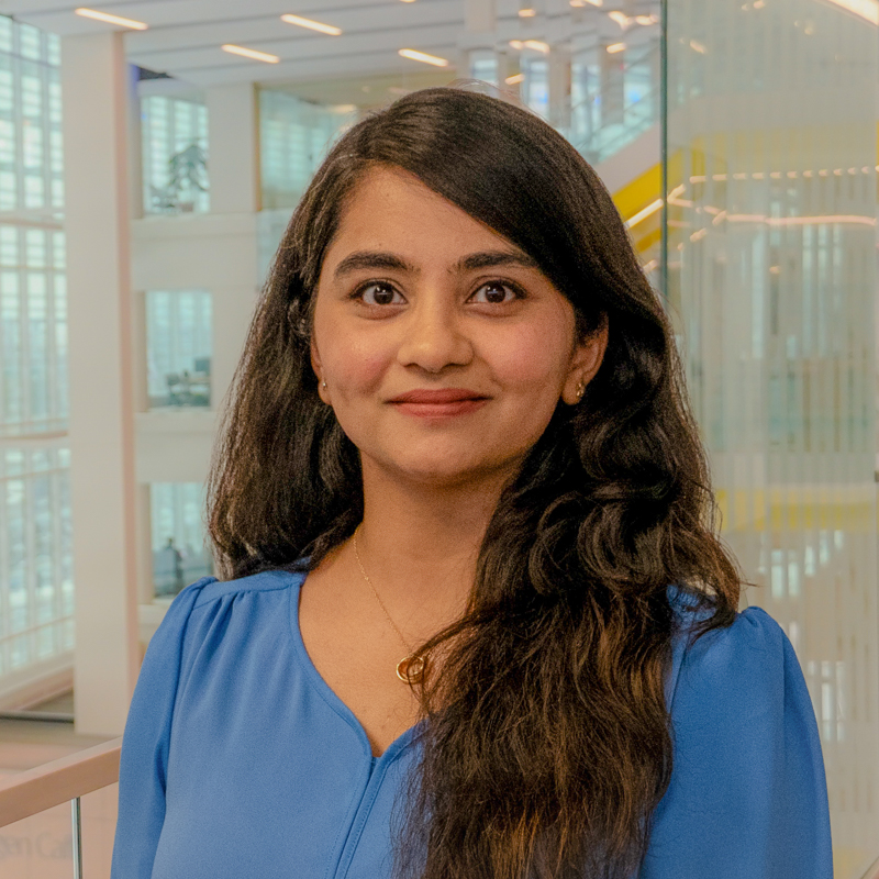 Headshot of Akhila Joshi smiling at the camera, wearing a blue blouse, with long dark hair and a bright, modern university building interior in the background.