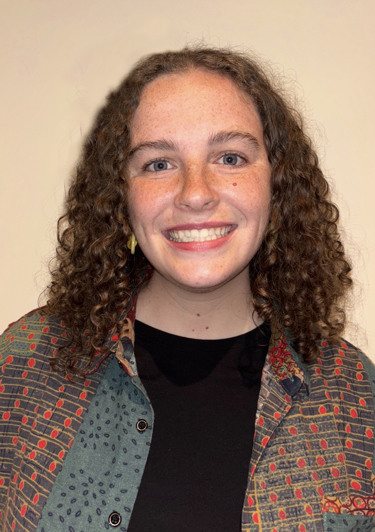 Photo of Ellie Seehorn, a PhD student in IDEA lab. They are white and present femininely—in this photo they have shoulder length brown curly hair and are wearing a colorful button down open over a black t-shirt, and are smiling